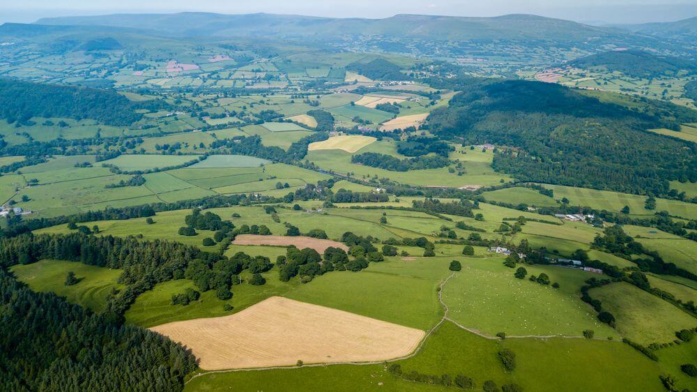 Aerial view of rolling Welsh countryside with patchwork farmland, forests, and distant mountains under clear blue sky.