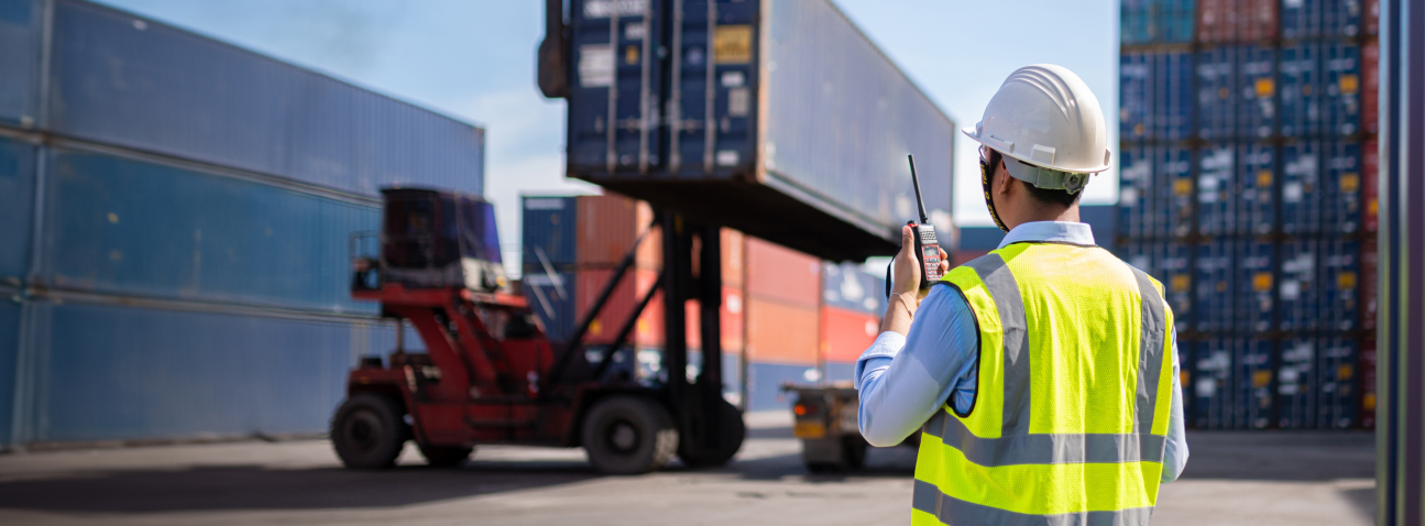 Worker in hard hat and safety vest communicating via radio at busy shipping container port with forklift and stacked cargo.