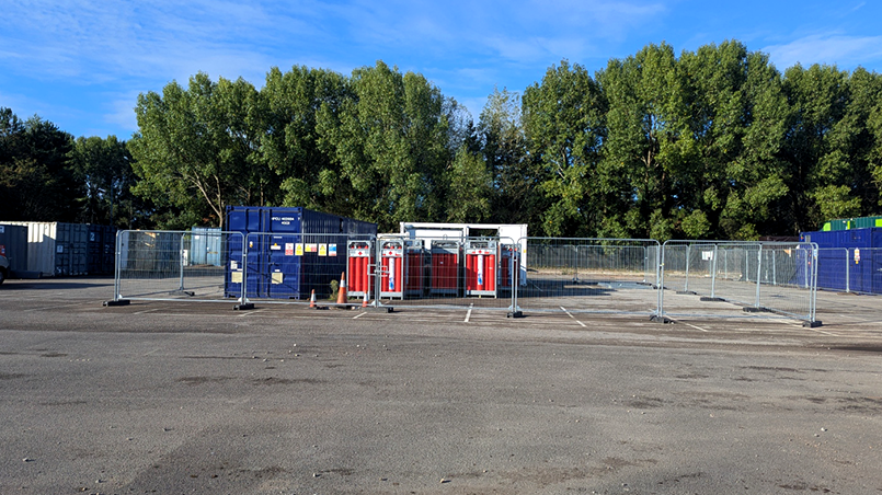 Construction site with blue shipping containers, red portable units, and metal fencing in South Wales, Pioneer 2 project.
